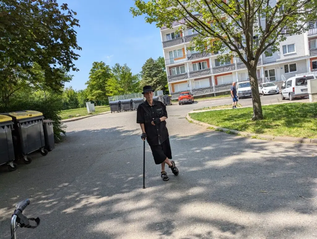 Timo-Thejus auf Asphaltstraße mit Wohnhaus und parkenden Fahrzeugen im Hintergrund.